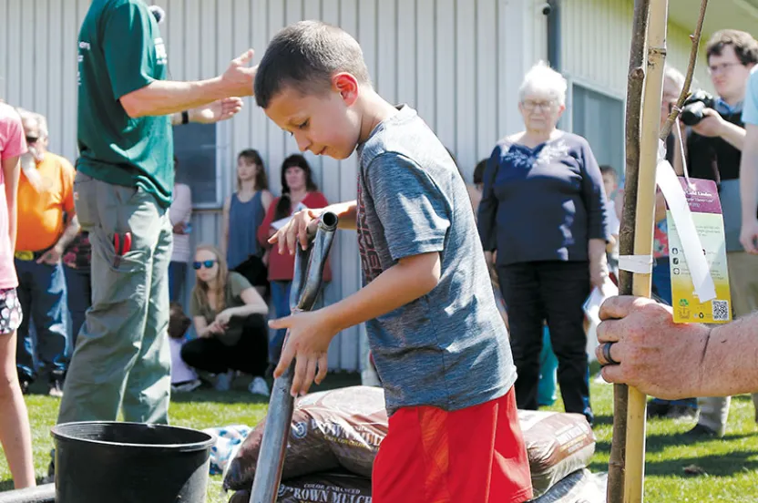 A young boy in a gray shirt and red shorts holds a gardening tool amid a crowd at a planting event near a building.