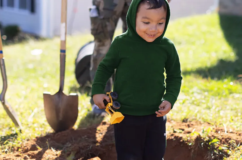 A child in a green hoodie holds a toy truck while standing near a freshly dug hole in the yard, with gardening tools in the background.