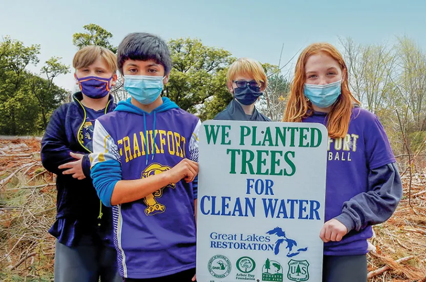A group of four children stands proudly beside a sign reading "We planted trees for clean water," surrounded by a wooded area.