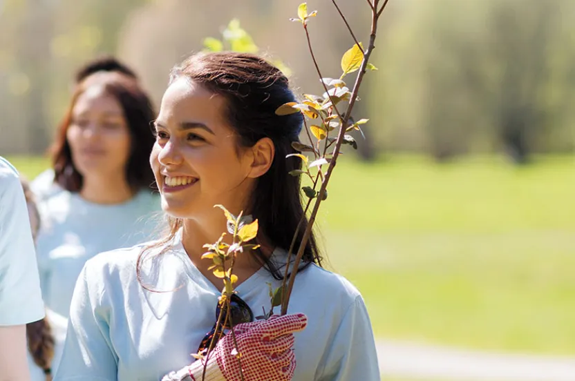 A group of volunteers in light blue t-shirts participate in a tree planting event, one holding young saplings outdoors.