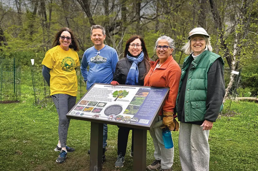 A group of five people stands in front of an informational sign about ecological restoration in a lush green area.