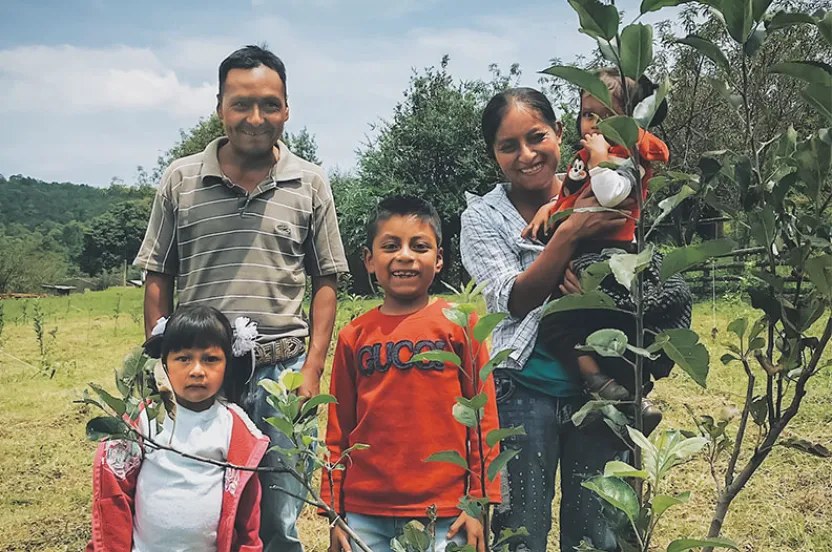 A group of children and adults stand together in a lush green field surrounded by young plants under a cloudy sky.