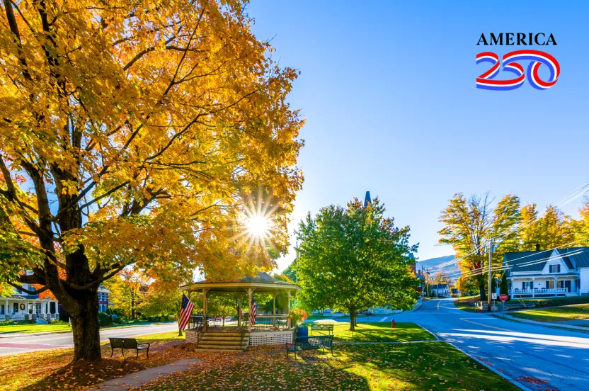 A scenic autumn street featuring vibrant yellow trees, a gazebo, benches, and American flags, with clear blue skies above.