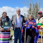 Four people outdoors on a sunny day, two holding shovels, one holding a "Tree City USA" plaque, wearing colorful traditional and modern clothing.